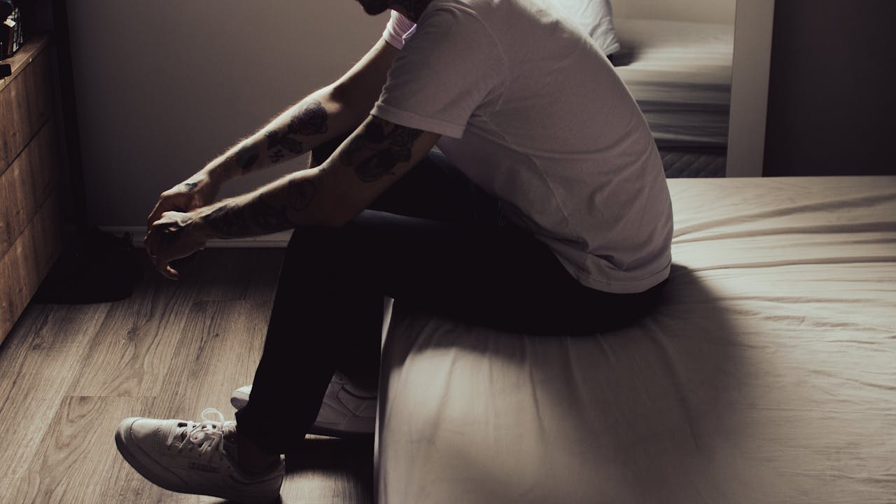 hero-img-02 A man sits thoughtfully on the edge of a bed in a dimly lit bedroom, evoking introspection.