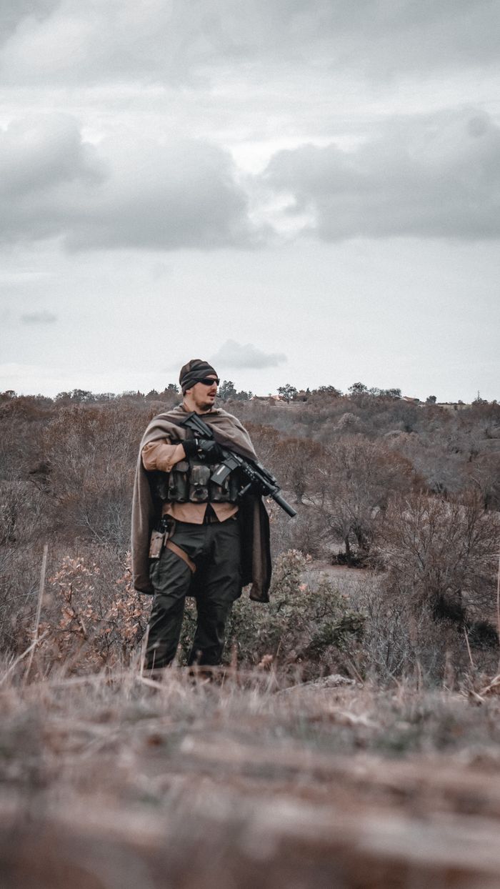 why-choose-us A soldier with a rifle standing in an outdoor field with overcast skies and rugged terrain.
